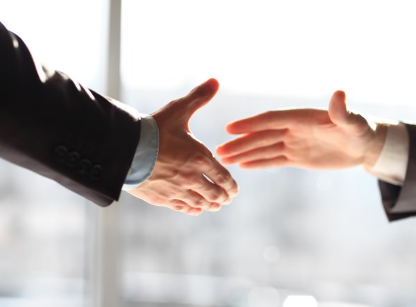 Zwei Menschen strecken ihre Hände für einen Handschlag aus. Beide tragen Anzüge, das Bild ist im Gegenlicht aufgenommen.